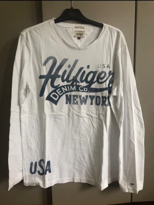 Vit långärmad topp Hilfiger Denim - Vit långärmad topp från Hilfiger Denim med stort blått tryck framtill där det står 'Hilfiger Denim Co. New York USA'. T-shirten har v-ringning och är gjord i mjuk bomull, perfekt för en avslappnad och sportig look.
