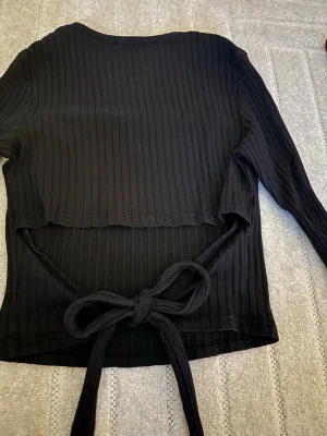 Svart ribbad topp med knytband - Svart långärmad topp med ribbad struktur och knytband i midjan. Toppen har rund halsringning och är figurnära i modellen. Perfekt för en trendig och stilren look.