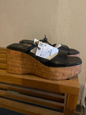 Svarta platåsandaler från ZARA - Säljer ett par svarta platåsandaler från ZARA med breda remmar i konstläder och cool korksula. Sandalerna har öppen tå och en smal rem med metallspänne över foten. Perfekta för dig som vill ha extra höjd och en trendig look.