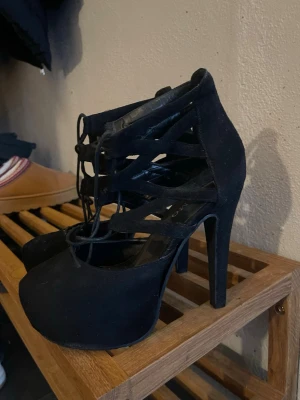 Svarta pumps med snörning fr Scorett - Säljer ett par svarta pumps med hög klack och snygga snörningsdetaljer över foten. Skorna har rundad tå och är tillverkade i mockaimitation som ger en lyxig känsla. Perfekta för dig som vill ha en edgy och trendig look.