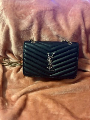 Svart YSL handväska i läder - Säljer en svart quiltad handväska från Saint Laurent med silverfärgad YSL-logga framtill och kedjerem i metall. Väskan har en klassisk rektangulär form och är tillverkad i slätt läder med snyggt mönster. Perfekt accessoar för att lyfta din outfit. Vet ej om den är äkta köptes av en annan tjej på plick.