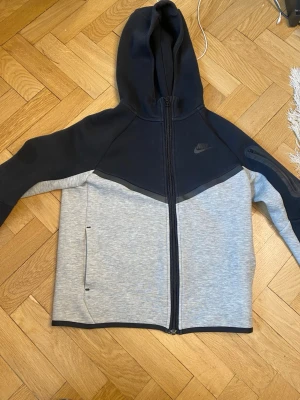 Nike hoodie i svart och grå med dragkedja - Nike hoodie i svart och ljusgrått med hel dragkedja framtill och två sidofickor. Hoodien har en stor huva och Nike-logga på bröstet. Tillverkad i mjukt sweatshirtmaterial, perfekt för en sportig och avslappnad stil.