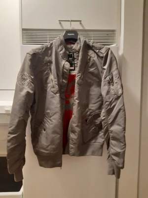 Grå bomberjacka Alpha Industries Falcon ll - Snygg grå bomberjacka från Alpha Industries modellen är Falcon ll med flera dragkedjeförsedda fickor, ribbade muddar och detaljer på ärmarna. Klassisk MA-1 stil med patch på ena ärmen och orange innerfoder. Perfekt för dig som vill ha en streetwear-look.