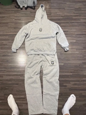 Grått mjukisset med luva - Säljer ett grått mjukisset med hoodie och matchande byxor. Setet har en broderad patch med svart, vitt och gult märke på både tröjan och byxan. Hoodien har magficka och muddar vid ärmslut. Perfekt för chill dagar hemma.