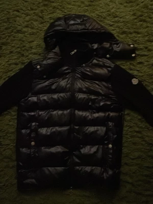 Svart dunväst med huva från Moncler - Snygg svart dunväst från Moncler med avtagbar huva och blanka paneler. Litet slitage som man ser på 4 bilden. Liten i storleken så M passar den