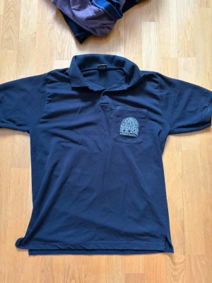 Marinblå pikétröja  - Marinblå pikétröja med krage och korta ärmar från IIM Ahmedabad. Bröstficka med vit broderad logga och text. Klassisk passform och mjukt material som känns skönt mot huden.