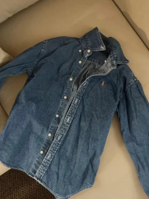 Blå denimskjorta från Ralph Lauren - Klassisk blå jeansskjorta från Ralph Lauren med knappstängning och ikonisk broderad logga på bröstet. Skjortan har lång ärm och en avslappnad passform. Perfekt att styla lager på lager eller bära som den är. Passar bättre S och Xs
