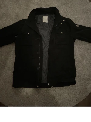 Svart jeansjacka med bröstfickor - Stilren svart jeansjacka med klassisk krage och två bröstfickor med lock och knapp. Jackan har knappstängning framtill och en diskret patch på ena ärmen. Perfekt för dig som gillar en enkel men cool look.