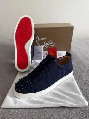 Christian Louboutin Skor - Snygga mörkblå sneakers från Christian Louboutin med klassisk röd sula och svarta nitar på tån. Skorna har snörning, vit platt sula och är tillverkade i mocka med läderdetaljer på insidan. Perfekta för dig som vill sticka ut med en exklusiv touch.