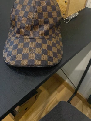 Louis Vuitton brunrutig keps - Säljer en snygg keps från Louis Vuitton med det klassiska bruna damier-mönstret. Kepsen har böjd skärm och är tillverkad i ett tåligt material med exklusiv känsla. Perfekt accessoar för dig som gillar lyxiga detaljer och streetstyle.