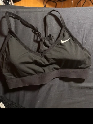 Svart sport-bh från Nike strl S - Svart sport-bh från Nike med smala axelband och inlägg med klassisk logga framtill.  Tillverkad i Dri-FIT-material som transporterar bort fukt och håller dig torr. Mjuk resår under bysten för extra stöd och komfort. Perfekt för träning eller vardagsbruk.