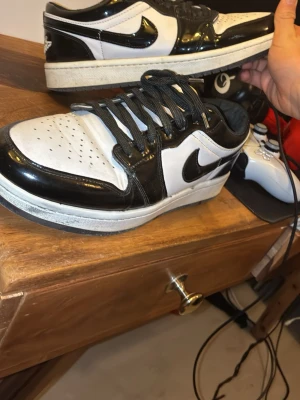 Nike Air Jordan 1 Low svart/vit - Snygga Nike Air Jordan 1 Low sneakers i svart och vitt med glansig finish. Skorna har klassisk låg siluett, svart swoosh och vita partier i läder. Perfekta för dig som gillar streetwear och vill ha en ikonisk look.