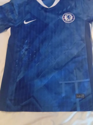 Chelsea tröja - Chelsea FCs officiella blå matchtröja från Nike med klubbmärke på bröstet och vit V-ringad krage. Tillverkad i lätt och ventilerande material, perfekt för fotbollsträning eller match. Snyggt mönster i olika blå nyanser och diskreta ränder.