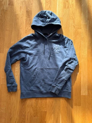 Blå hoodie från Dope - Snygg blå hoodie från Dope med stor vit logga på ryggen och mindre tryck på bröstet. Klassisk känguruficka framtill, justerbar huva med snören och ribbade muddar. Perfekt för en avslappnad streetwear-look.