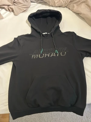 Svart hoodie från Antony Morato - Snygg svart hoodie från Antony Morato med stor logga framtill i metallic- och gröna detaljer. Klassisk känguruficka och justerbar huva med snören.