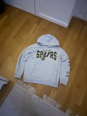 Grå San Antonio Spurs hoodie H&M - Snygg grå hoodie från H&M med San Antonio Spurs-logga i svart och guld på bröstet och tryck på ena ärmen. Klassisk känguruficka framtill och mjukt material som känns skönt mot huden. Perfekt för dig som gillar basket och streetwear.