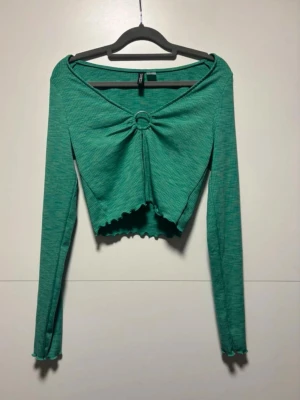 Grön långärmad crop top med ringdetalj - Trendig grön långärmad crop top med ringdetalj framtill och vågig kant nertill. Toppen har en djup v-ringning och är tillverkad i ett mjukt, ribbat material. Perfekt för dig som vill ha en cool och unik look.