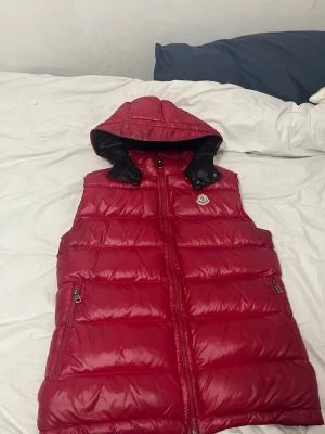 Röd dunväst från Moncler med huva - Snygg röd dunväst från Moncler med avtagbar huva och dragkedja framtill. Västen har två fickor med dragkedja och klassisk Moncler-logga på bröstet. Perfekt för lager-på-lager och riktigt skön att bära under kyliga dagar.