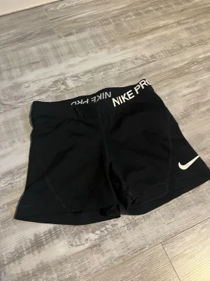 Svarta Nike Pro träningsshorts - Säljer ett par svarta Nike Pro träningsshorts med vit logga och bred resår i midjan där det står 'NIKE PRO'. Perfekta för gym, löpning eller annan träning. Snygg och sportig design i stretchigt material.