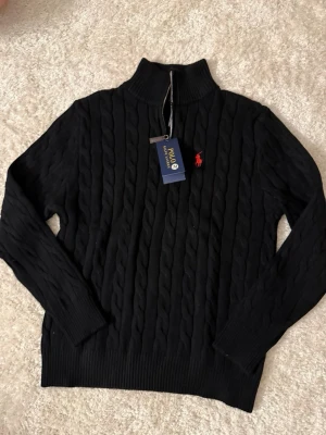 Svart kabelstickad tröja Polo Ralph Lauren - Kabelstickad svart tröja från Polo Ralph Lauren med halv dragkedja och hög krage. Tröjan har det klassiska röda Polo-logot broderat på bröstet och ribbade muddar vid ärmslut och nederkant. Perfekt för en stilren och avslappnad look. Det står storlek M men skulle nog säga den passar bättre S/Xs.