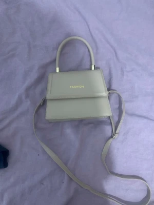 Beige minihandväska med axelrem - Snygg beige minihandväska  med både handtag och justerbar axelrem. Väskan har en stilren, fyrkantig form och guldiga detaljer vid handtaget. Perfekt för dig som gillar minimalistisk och trendig design.