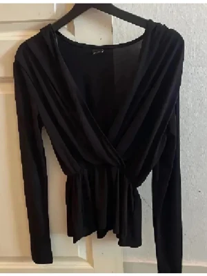 Svart omlottopp med lång ärm - Snygg svart omlottopp med lång ärm och markerad midja. Toppen har djup v-ringning och är tillverkad i ett mjukt, stretchigt material som ger en smickrande passform. Perfekt att styla med jeans eller kjol för en trendig look.