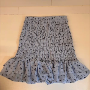 Ljusblå blommig volangkjol - Supersöt ljusblå kortkjol med små svarta blommor och volangkant. Storlek 158