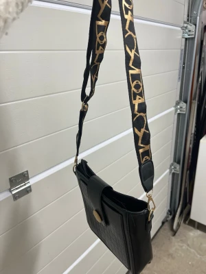 Svart axelväska med bred rem - Cool svart axelväska med bred, justerbar rem med guldmönster. Väskan har en stilren, rektangulär form och ett lock med knappstängning. Materialet ser ut som konstläder med en lätt struktur. Perfekt för att bära det viktigaste. Inte använd nå mycket! 