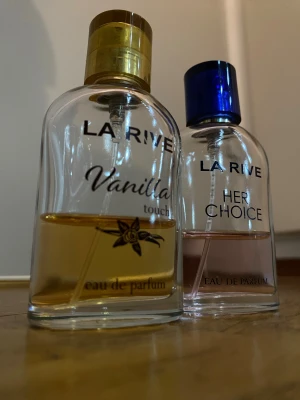 La Rive Vanilla Touch & Her Choice parfym - Två flaskor eau de parfum från La Rive: Vanilla Touch med guldigt lock och gul vätska, samt Her Choice med blått lock och rosa vätska. Båda flaskorna är transparenta och har rundad form. Perfekt för dig som gillar söta och fräscha dofter.