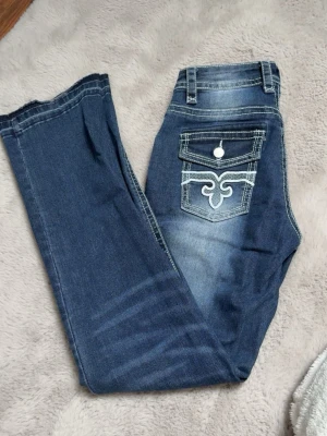 Blå jeans med tryck på bakficka - Snygga blå jeans med raka ben, Bakfickan har ett vitt broderat mönster och knappdetalj. 