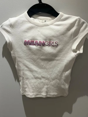 Vit Mean Girls t-shirt från H&M - Säljer en vit ribbad t-shirt från H&M med rosa texten 'MEAN GIRLS' på bröstet. T-shirten är kortärmad och har en croppad passform som ger en trendig look. Perfekt för dig som älskar popkultur.