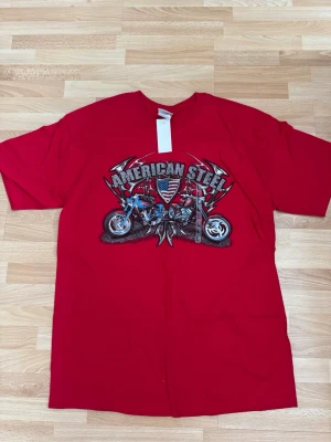 Ny. Röd American Steel t-shirt - Cool röd t-shirt med tryck av två motorcyklar och texten 'American Steel' på bröstet. Sköld med amerikanska flaggan i mitten och detaljer i silver och blått. Klassisk passform och rund hals, perfekt för dig som gillar bikerstil.