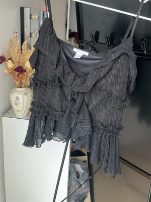 Svart volangig linnetopp från Amisu - Säljer en svart linnetopp från Amisu med tunna axelband, volangdetaljer och ribbad struktur. Toppen har flera lager volanger och en luftig, croppad passform som ger en cool och trendig look.