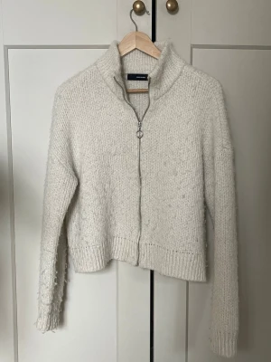 Vit stickad ziptröja från Vero Moda - Mysig vit stickad tröja från Vero Moda med dragkedja framtill och hög krage. Tröjan har lång ärm och ribbade muddar vid ärmslut och nederkant. Perfekt för lager på lager och enkel att matcha med jeans eller kjol.