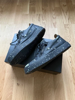 Svarta skor från Giuseppe Zanotti  - Unika svarta sneakers från Giuseppe Zanotti med vit reptilmönstrad ovandel, svarta snören och dragkedjor på sidorna. Skorna har svart sula med vita färgstänk. Box och påse följer med till skorna och de är bara använda en gång