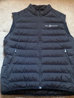 Sail racing väst - Snygg svart dunväst från Sail Racing med quiltad design och två dragkedjeförsedda fickor framtill. Västen har hög krage och logga på bröstet. Perfekt för lager-på-lager och håller dig varm under kyliga dagar. Inte använd 