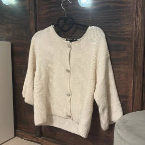 Krämvit kofta från Gina Tricot - Mysig krämvit kofta från Gina Tricot med dekorativa silvriga/diamant knappar. Koftan har rund halsringning, ribbstickade muddar och en avslappnad passform. Perfekt att slänga på sig när det är kyligt ute. Väl använd men fortfarande väldigt bra skick. Lite fläckar runt halsringen men inte synligt längre ifrån!