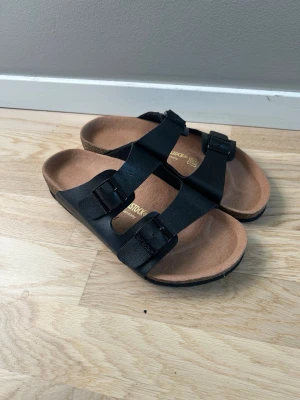 Birkenstock Arizona  - Helt nya endast prövade en gång Klassiska svarta sandaler från Birkenstock med två breda remmar och justerbara svarta spännen. Fotbädden är i kork och mocka för extra komfort. Perfekta för en avslappnad och stilren look.