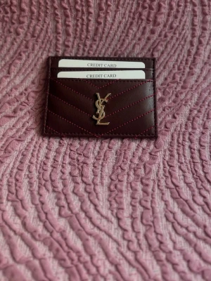 Vinrött kortfodral från Saint Laurent - Säljer among ett stil firm vinrött kortfodral från Saint Laurent   med ikoniska YSL son i guld framtill. Fodralet har flera kort sig ten och är gjort i slätt läder that har dekor 'stickade' sömmar i rött. Perfekt för dig som vill ha något lyxigt och smidigt i väskan.