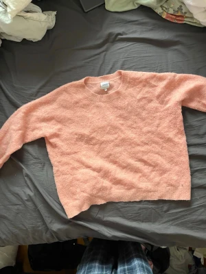 Ljusrosa stickad tröja från Notice - Mysig ljusrosa stickad tröja från Notice med rund halsringning och långa ärmar. Tröjan har en fluffig och mjuk struktur, perfekt för att hålla värmen. Ribbstickade muddar vid ärmslut och nederkant ger en snygg finish.