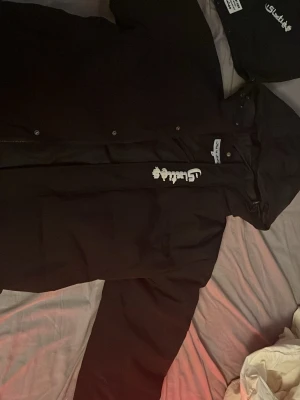 Svart vinterjacka från sikties  - Svart vindjacka från Sikties med avtagbar huva och tryckta vita loggor på bröstet och huvan. Jackan har dragkedja och tryckknappar framtill samt justerbar huva. Materialet är lätt och vindtätt, perfekt för blåsiga dagar.har använt några gånger bara 