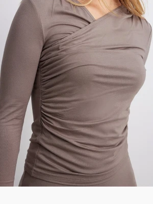 Taupe omlottopp med rynkade detaljer - Taupefärgad omlottopp med lång ärm och rynkade detaljer längs sidorna. Toppens material är stretchigt och sitter tight mot kroppen, vilket ger en smickrande passform. Perfekt att styla med jeans eller kjol för en trendig look. Endast använd fåtal gånger