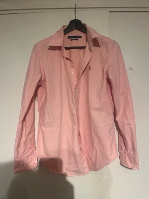 Rosa skjorta från Ralph Lauren - Snygg rosa skjorta från Ralph Lauren i super slim fit-modell. Skjortan har klassisk krage, långa ärmar och knäppning framtill. Perfekt för dig som gillar en stilren och fräsch look.