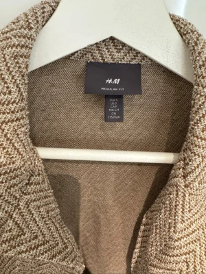 Beige kortärmad skjorta från H&M - Kortärmad skjorta från H&M i beige med fiskbensmönster. Skjortan har klassisk krage och regular fit, perfekt för en avslappnad men ändå stilren look. Materialet känns mjukt och har en lätt struktur.