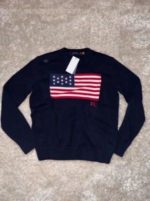 Mörkblå stickad tröja Polo Ralph Lauren - Snygg mörkblå stickad tröja från Polo Ralph Lauren med amerikansk flagga i rött, vitt och blått på bröstet samt RL-broderi. Tröjan har rund halsringning och ribbstickade muddar. Perfekt för dig som vill ha en klassisk och trendig look.