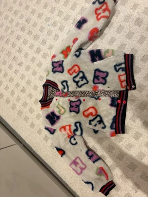 Färgglad vändbar bomberjacka/fuskpäls/barn - Säljer en unik vändbar bomberjacka med ena sidan i vit fluffig teddy med färgglada bokstäver och andra sidan i leopardmönster. Jackan har ribbade muddar i svart, rött och rosa samt knappar framtill. Perfekt för dig som vill sticka ut och variera din stil.