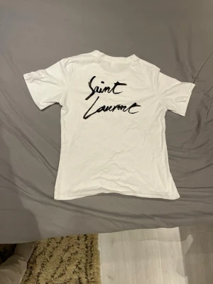 Vit Saint Laurent t-shirt - Vit t-shirt från Saint Laurent med svart logotyptryck både fram och bak. Klassisk passform med rund hals och korta ärmar. Tillverkad i mjuk bomull som känns skön mot huden. Perfekt för dig som gillar stilrena och ikoniska plagg. Storlek S.  Sparsamt använd.