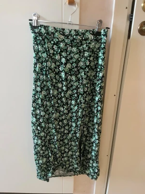 Grön blommig långkjol med slits - Snygg långkjol med grönt och vitt blommönster på svart botten. Kjolen har en slits framtill som ger en trendig touch. Perfekt för dig som gillar en feminin och avslappnad stil. Materialet känns lätt och luftigt. Om man vill ha fler bilder på plagget (på eller andra vinklar) är det bara att säga till så fixar jag det