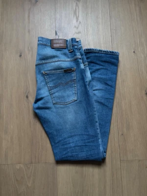 Blå raka jeans från Lee - Säljer ett par klassiska blå jeans från Lee med rak passform och fem fickor. Jeansen har en snygg tvättad look, bruna detaljer vid linningen och är tillverkade i slitstarkt denim. Perfekta för en avslappnad stil.
