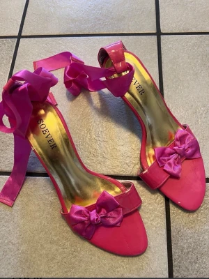 Rosa pumps med rosett från Soever - Snygga rosa pumps från Soever med spetsig tå, smal klack och dekorativa rosetter framtill. Skorna har breda satinsnören som knyts runt ankeln för en extra statement-look. Perfekta för dig som vill sticka ut med färg och detaljer.